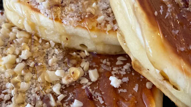 A close-up of pancakes in syrup, with crushed nuts and coconut flakes scattered on top