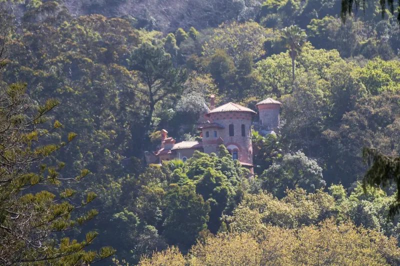 A stone house with a rounded reddish tower rising out of dense hillside forest