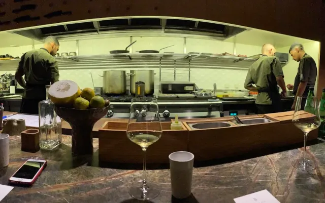 An open kitchen with three cooks, a marble counter and wine glasses in the foreground