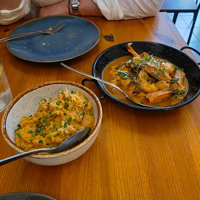 A bowl of orange mash with spring onion and a pan of shrimp in sauce on a wooden table