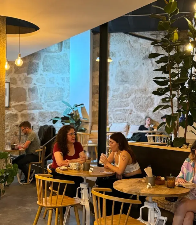 A space with stone walls, round tables, yellow wooden chairs and people sitting and chatting