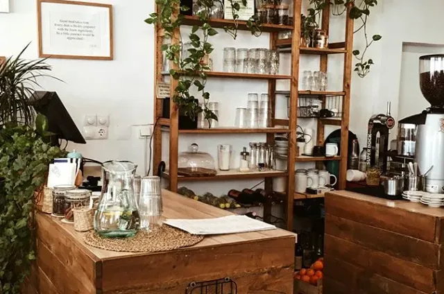 A wooden counter with a water jug and glasses, an open shelf unit with dishes, plants and a grinder
