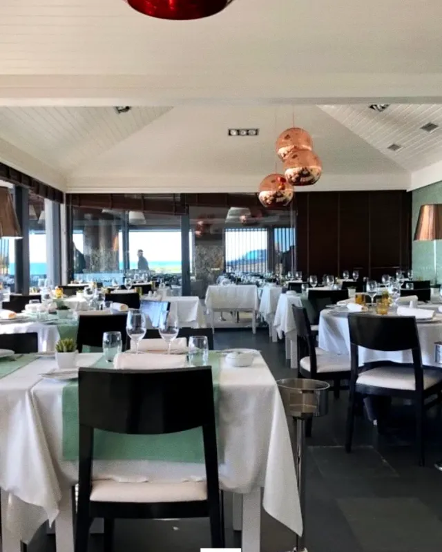 A room with white tablecloths, hanging copper lamps and large windows onto the sea