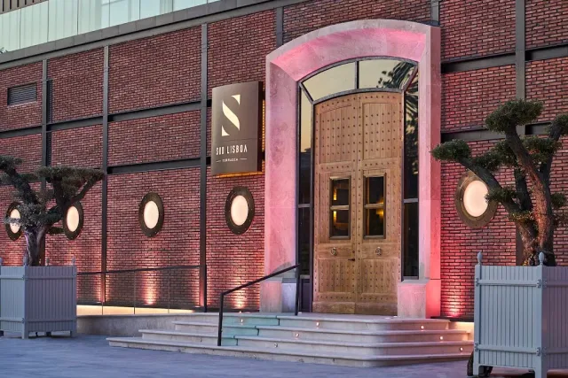 An arched wooden entrance in a brick wall lit pink, with trees in planters
