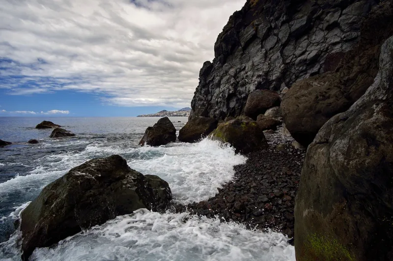 Waves breaking over a dark pebble shore at the foot of a high basalt cliff