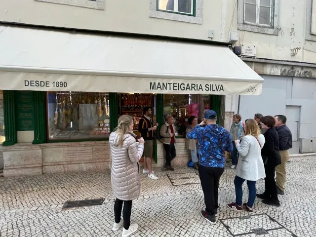 A corner shopfront with a cream awning, green frames, hanging cured meats in the window and a group of visitors