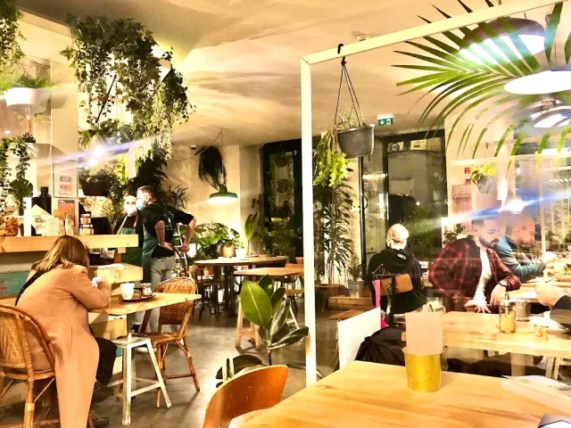A warm space crowded with hanging plants and pots, wooden tables and diners in yellowish light
