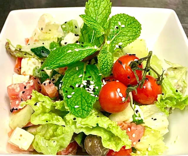 A white bowl with lettuce, cherry tomatoes, cheese cubes, sesame seeds and mint leaves