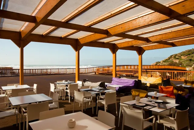 A wide terrace under a wooden pergola with white tables and beanbags facing a rough sea