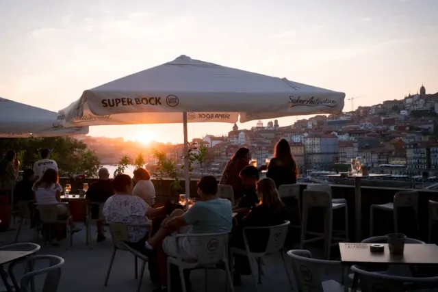 Diners under a parasol at sunset facing a riverbank and houses on a slope
