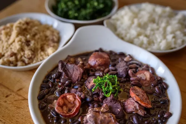 A bowl of black bean stew with sausage and meat, beside rice and toasted flour