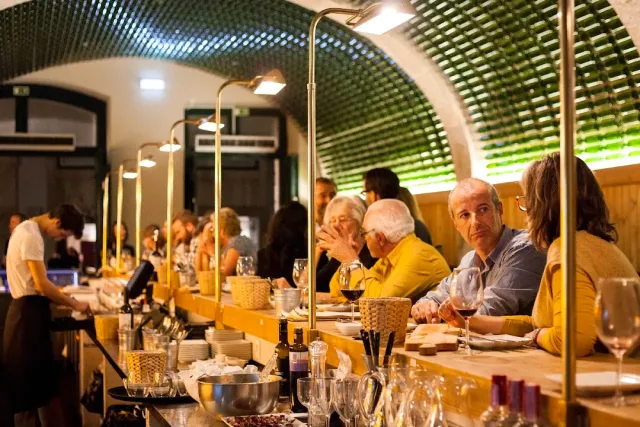A vaulted ceiling lined with green bottles above a long counter with diners