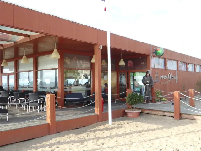 A reddish wooden building on the sand with a deck, chairs and a wide window wall