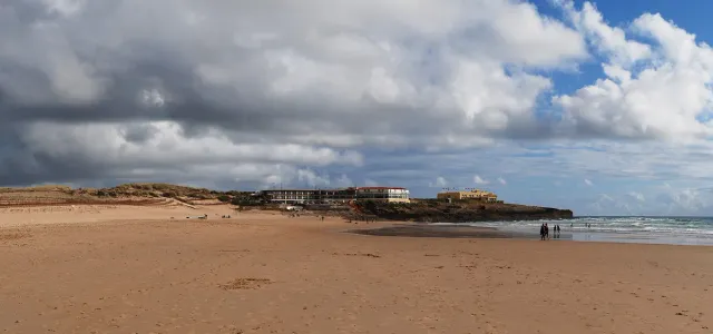 A wide sandy beach with dunes and a rocky headland under a cloudy sky