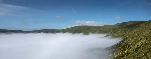 A green crater rim encircling a vast hollow filled with cloud