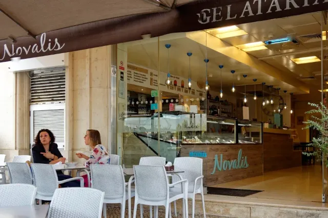 A gelato shop front with a chilled display case, white chairs outside and two seated women