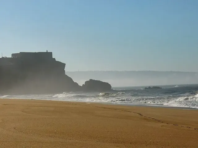 A wide sandy beach and white waves facing a cliff topped by a fort, in hazy air