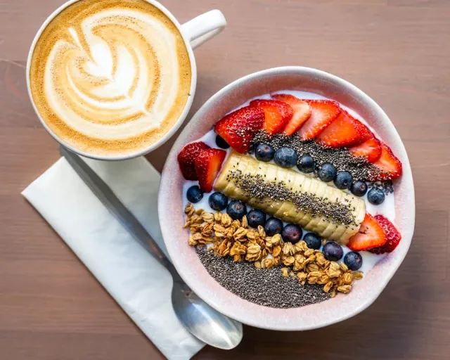 An overhead view of a cappuccino and a yoghurt bowl with strawberries, banana, blueberries and granola