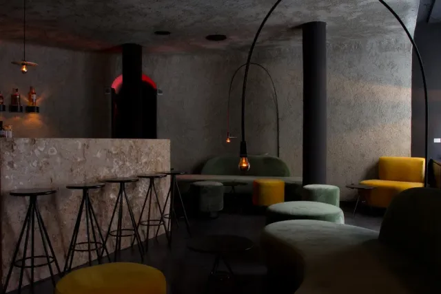 A dim concrete walled room with a stone counter, stools and yellow and green ottomans