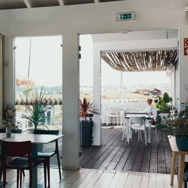 A bright white space with tables and an opening to a wooden deck shaded with reeds