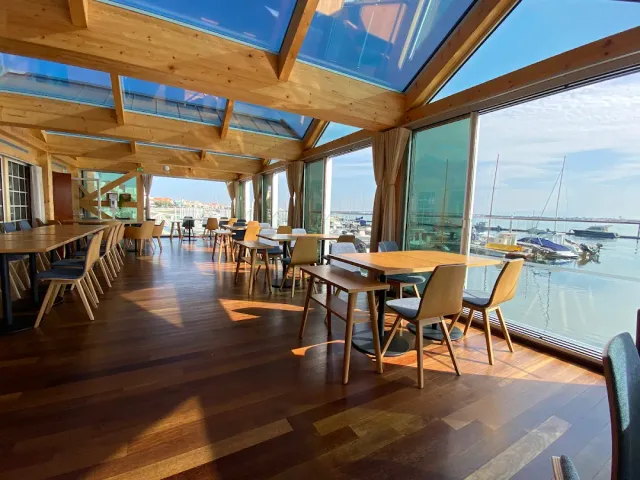 A room with wooden beams and a glass roof, light tables and windows onto a marina and boats