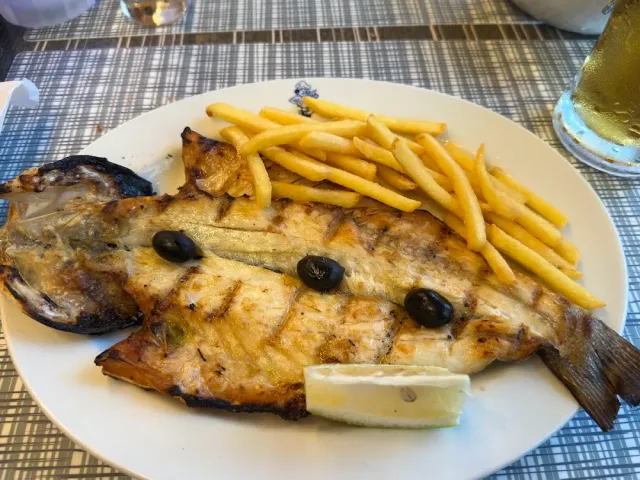 A whole grilled fish on a white plate beside fries, olives and a lemon wedge