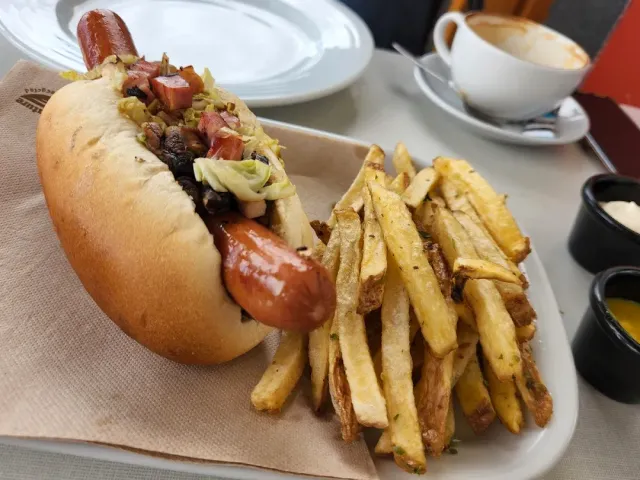 A hot dog in a bun with fries on a white plate beside a cappuccino cup