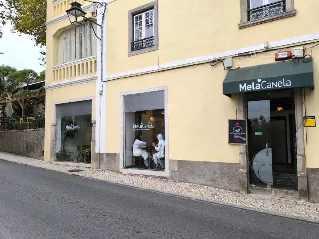 A yellow building on a sloping street with a dark green awning and two people sitting by the window