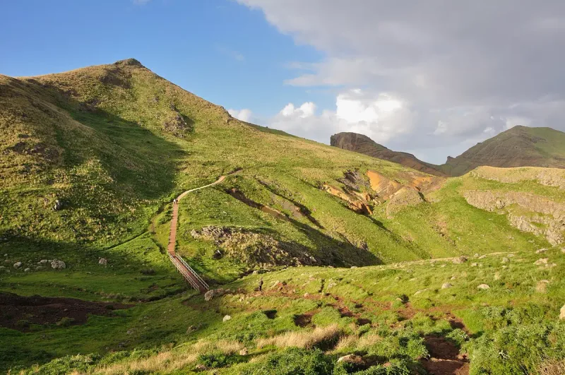 Footpath and wooden steps climbing a green ridge below rocky peaks and drifting cloud