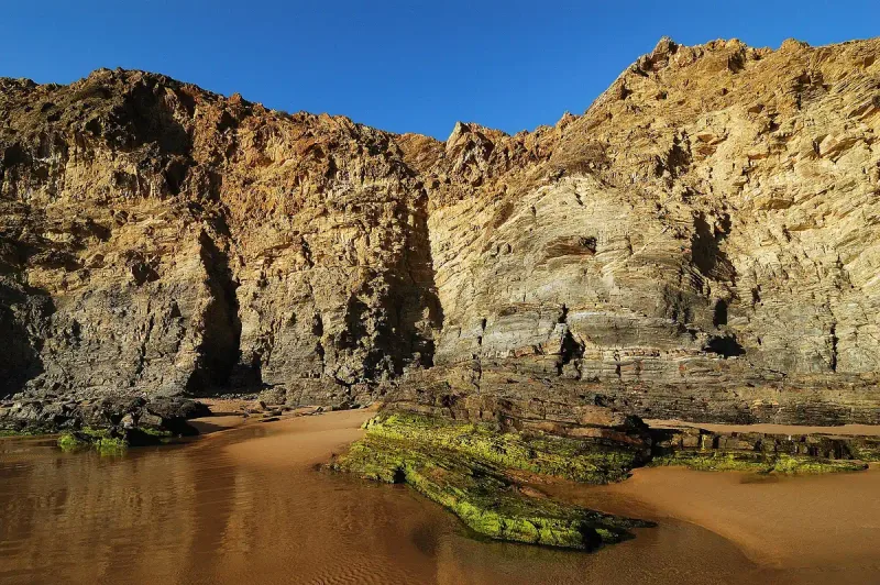 Tall rugged cliffs dropping to a strip of dark sand and rock pools