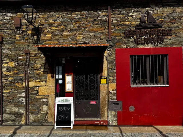 A dark stone facade with a red wall, a decorated iron door and a chalkboard sign at the entrance