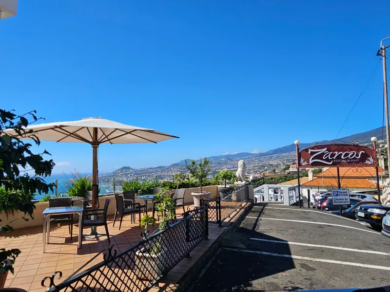 A terrace with tables and a parasol facing a bay and hillside town, beside the restaurant sign