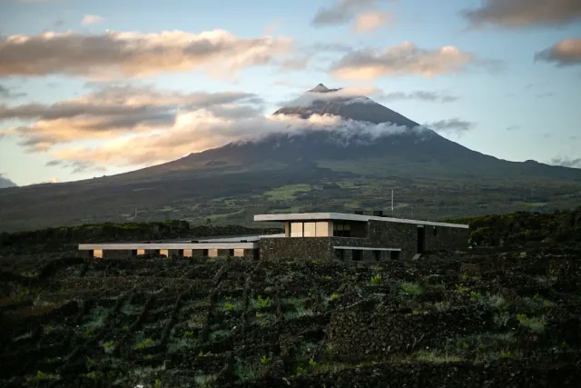 A low stone building on lava ground, with a cloud covered volcano behind it