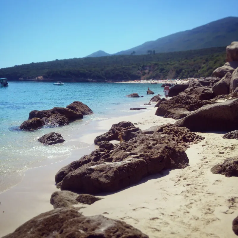 White sand and rounded rocks along clear turquoise shallows with a steep green hillside behind