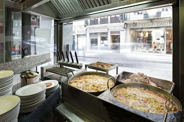 Large cooking trays on a hot counter facing a window onto the street, and stacks of plates