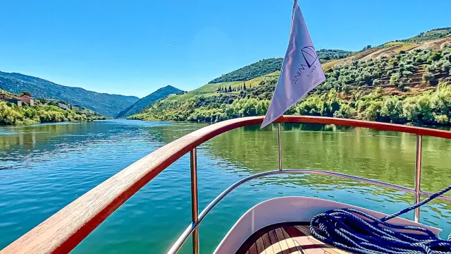The bow of a wooden boat with a pennant, a greenish river between terraced hills