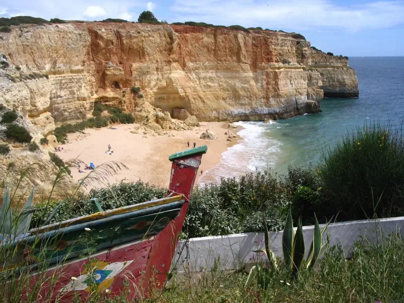 Small cove of golden sand and green water between reddish cliffs with a painted wooden boat in the foreground