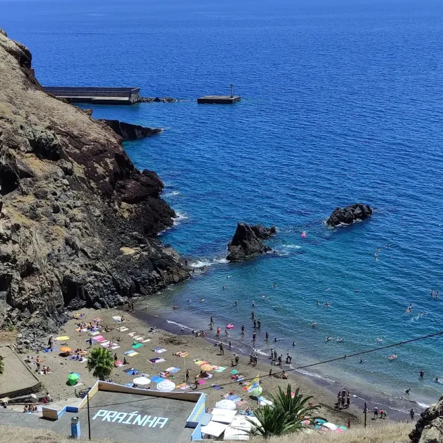 A dark sand bay with colorful umbrellas and bathers, beneath a reddish cliff