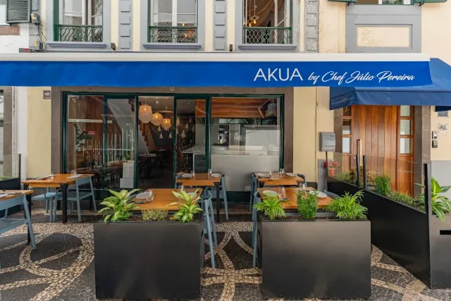 A facade with a blue awning, wooden tables outside and planters on a mosaic pavement