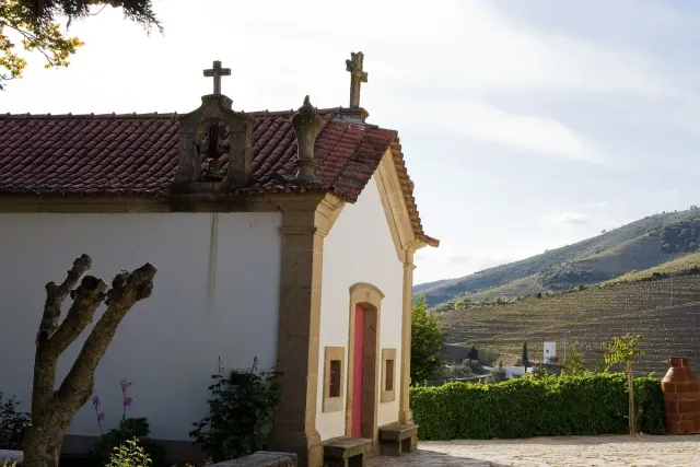 A white chapel with a tiled roof and crosses, and a vineyard hill behind