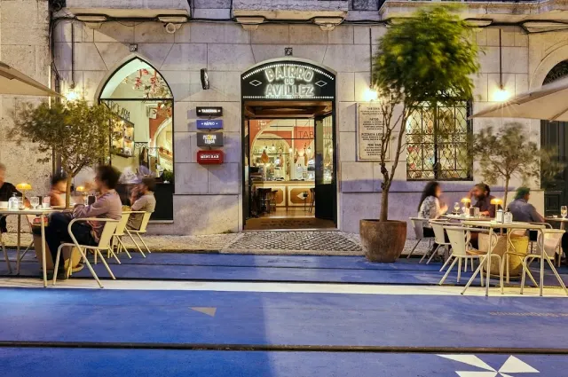 A stone facade with an arched entrance and lit pavement tables in the evening