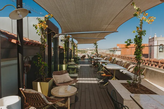 A rooftop terrace shaded by a canopy with sofas, planters and tables beside tiled roofs