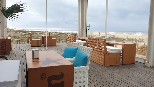 A wooden deck with sofas and turquoise cushions facing a sandy beach and grey sea