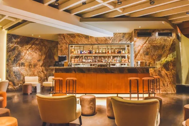 A wooden bar counter facing a brown marble wall, a bottle shelf and low armchairs