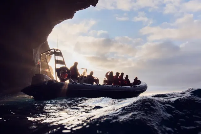 The silhouette of an inflatable boat with passengers by a dark cliff in low sunlight