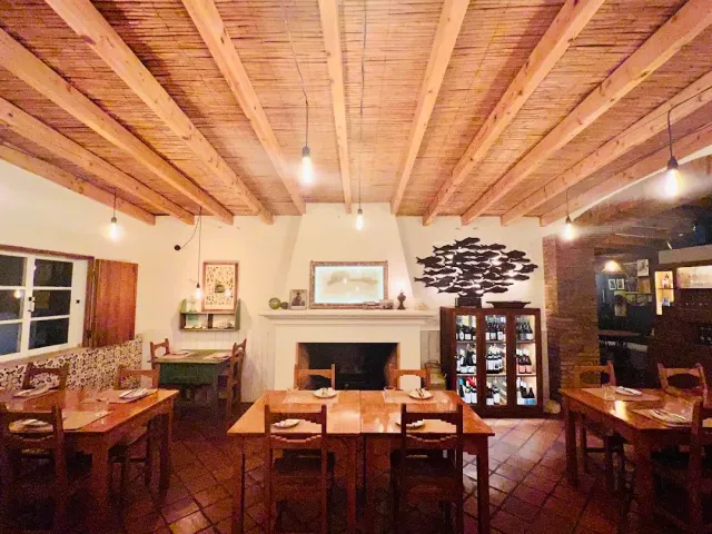 A room with a wooden beam ceiling, a white fireplace, a wine cabinet and wooden tables