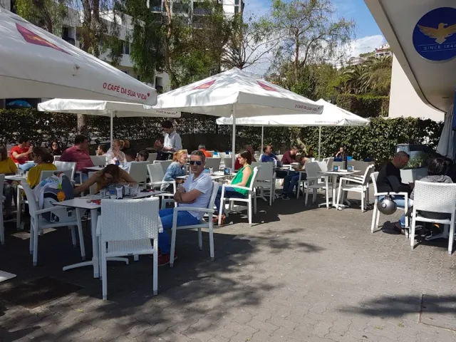 A terrace crowded with diners under white parasols, white plastic chairs and trees behind