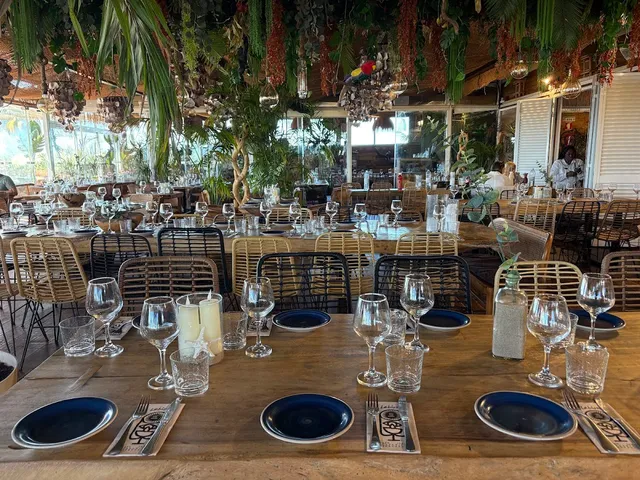 A hall of wooden tables set with wine glasses under a ceiling full of hanging plants