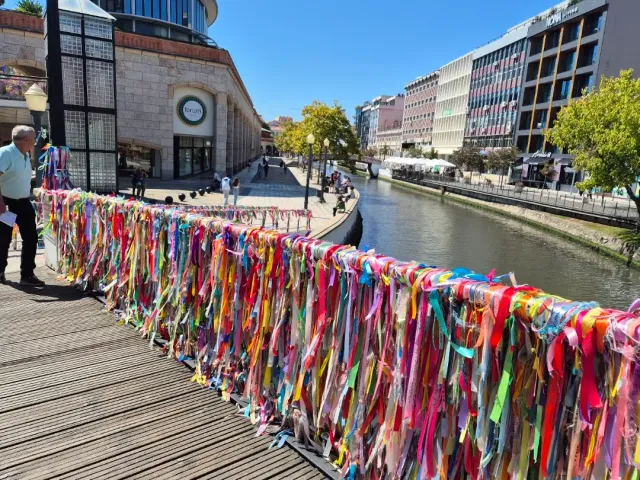 A bridge railing wrapped in thousands of colorful ribbons above a city canal