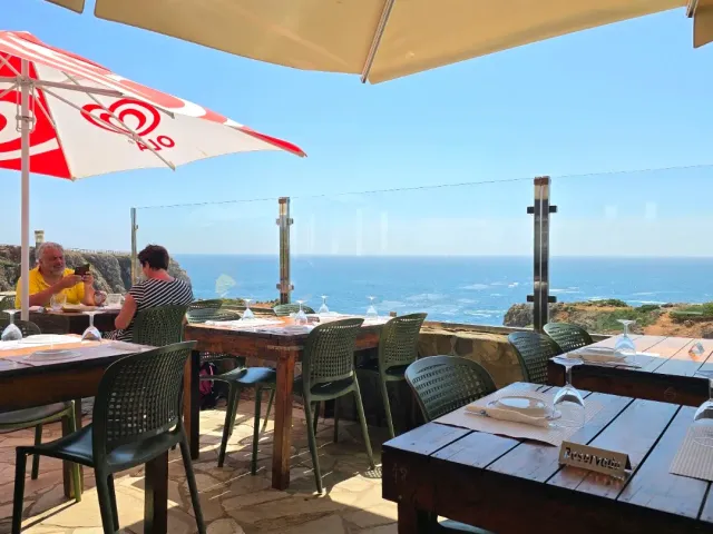 A terrace with parasols and green chairs above the ocean, diners sitting by the railing